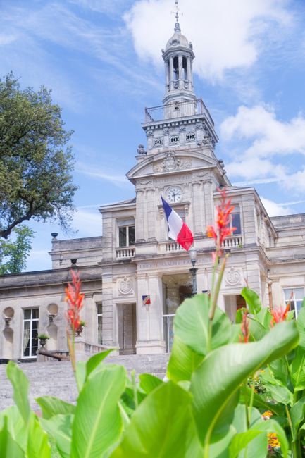 A close-up view of a vibrant floral arrangement featuring bright red ginger flowers and lush green leaves in the foreground, partially obscuring a historic building with a clock tower and a French fla