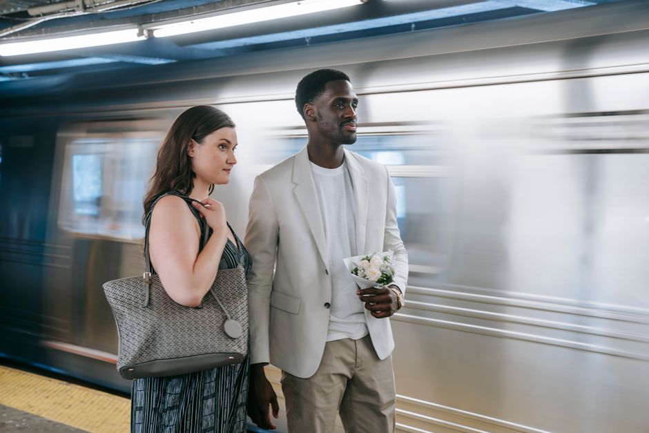 A woman in a dark striped dress holding a large grey patterned handbag stands next to a man dressed in a white blazer and light trousers, who is holding a small bouquet of white and pale pink flowers 