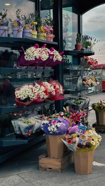 A display of fresh floral arrangements and bouquets arranged on dark grey shelves and wooden crates at a flower shop or outdoor market stall. The flowers include large white and pink chrysanthemums, v