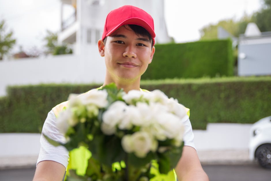 A delivery person wearing a bright red cap and a high-visibility vest presents a bouquet of fresh white roses and lisianthus, accented with green foliage, towards the camera in an outdoor suburban set