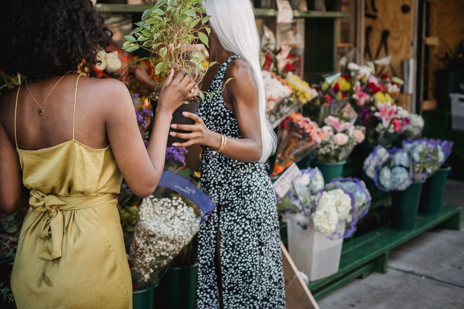 Two women are engaged in a conversation inside a flower shop, surrounded by an array of freshly cut floral arrangements. The woman on the left wears a sleeveless yellow dress with thin straps and a ti