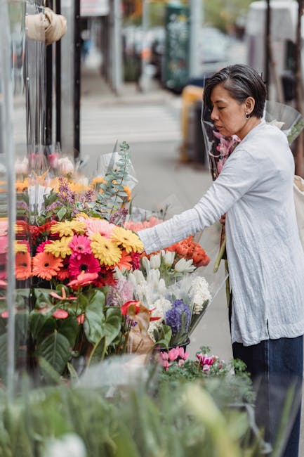 A woman with short dark hair, wearing a light grey cardigan, is selecting a vibrant bouquet of fresh flowers from an outdoor floral stall on Hornsey High Street. The arrangement includes bright yellow